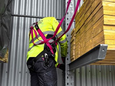 Person mit Schutzkleidung arbeitet an Metallregal mit Holzplanken, zeigt sicheren Umgang mit Baumaterialien.