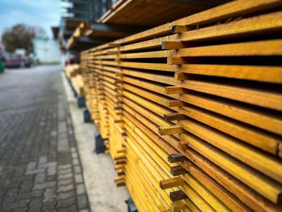 Gestapelte Holzbretter lagern draußen entlang gepflastertem Weg, zeigen geordnetes Baumaterial.